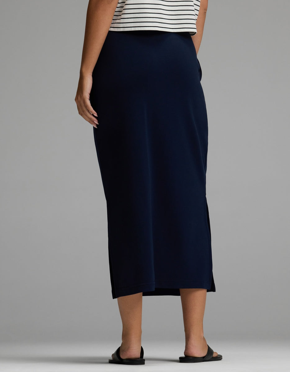 A person is shown from the back against a plain gray background, wearing the Cozy Earth Women’s Brushed Bamboo Midi Skirt in navy blue, paired with a striped top and black sandals.