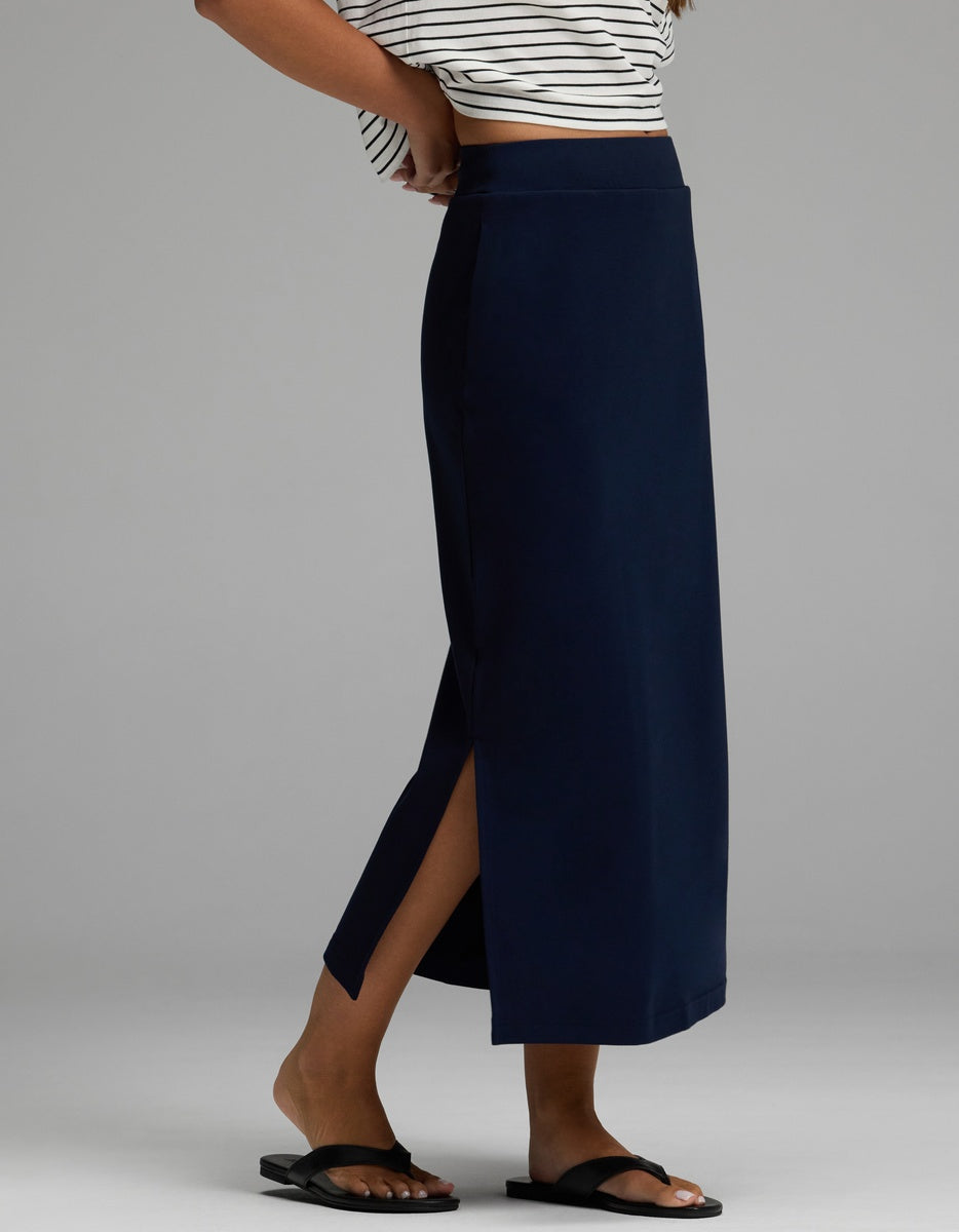 A person is wearing the Cozy Earth Women’s Brushed Bamboo Midi Skirt in navy blue with a side slit, paired with black slide sandals and a cropped striped top, standing against a plain gray background.