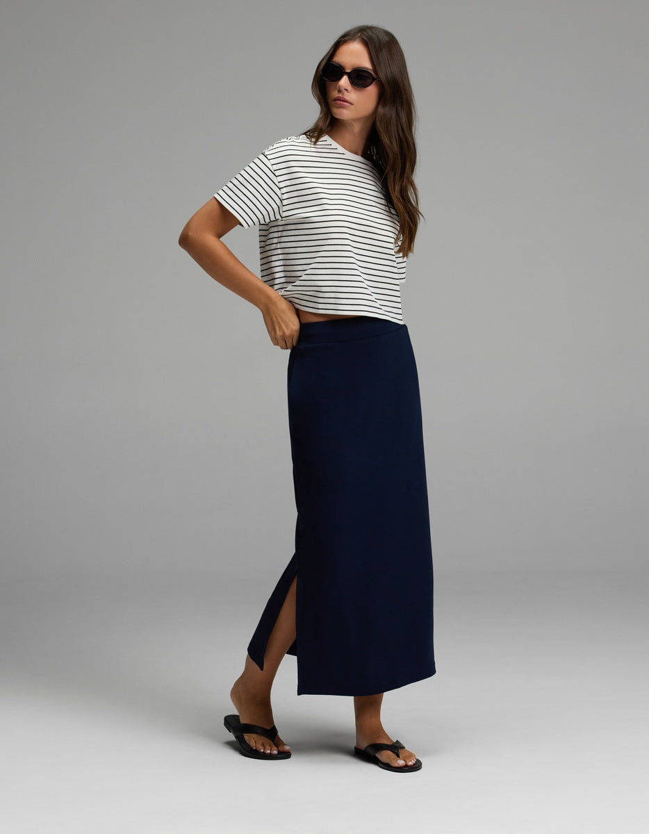 A woman wearing sunglasses, a white and black striped t-shirt, black sandals, and the Cozy Earth Women’s Brushed Bamboo Midi Skirt in navy blue with a side slit, standing against a plain gray background.