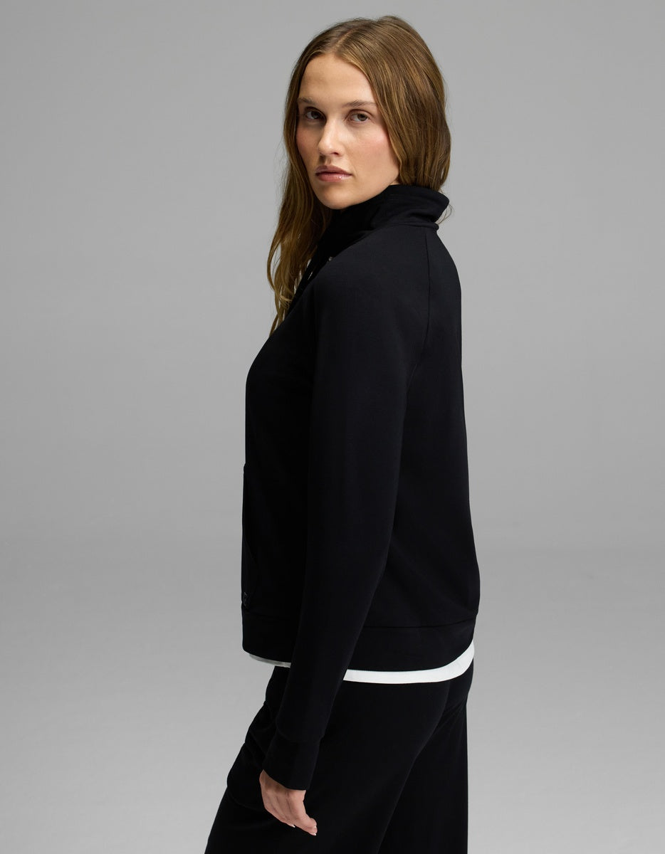 A woman with long, light brown hair stands sideways, looking at the camera. She is wearing the Cozy Earth Women’s Brushed Bamboo Mock Neck—a black zip-up jacket layered over a white top and paired with black pants—against a plain gray background.