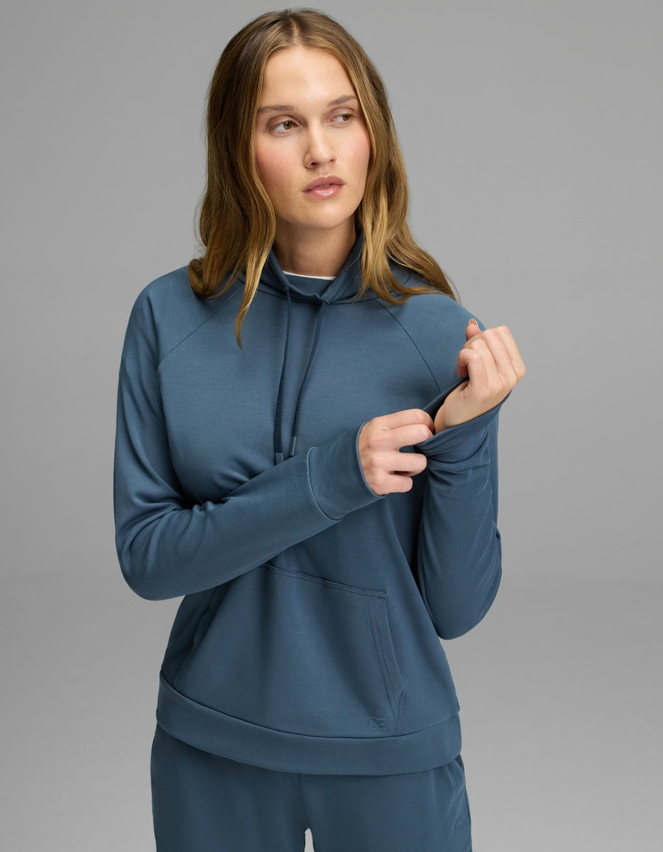 A woman wearing the Cozy Earth Women’s Brushed Bamboo Mock Neck in Dawn stands against a gray background, looking to the side while pulling at the sleeve’s thumb hole.|Color:Dawn