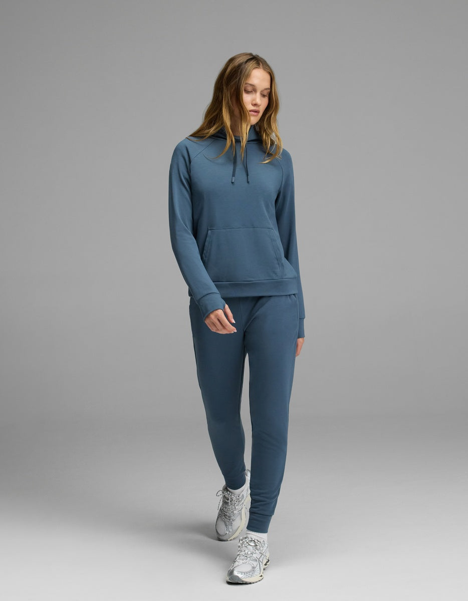 A person wearing the Cozy Earth Women’s Brushed Bamboo Mock Neck and matching joggers walks forward on a plain gray background, looking down with one hand in the top’s pocket and wearing white sneakers.