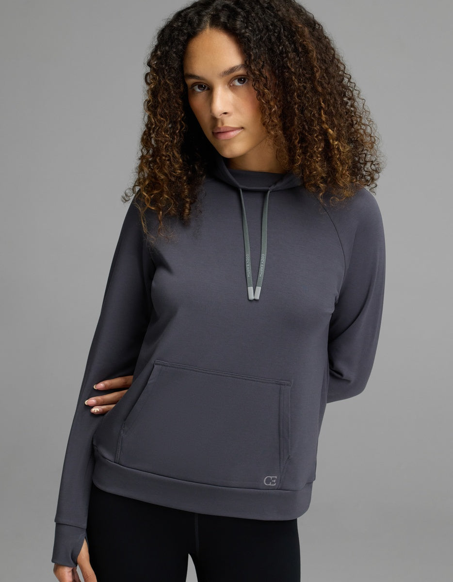 A woman with curly brown hair wears the Cozy Earth Women’s Brushed Bamboo Mock Neck in graphite, paired with black leggings, standing against a plain gray background.