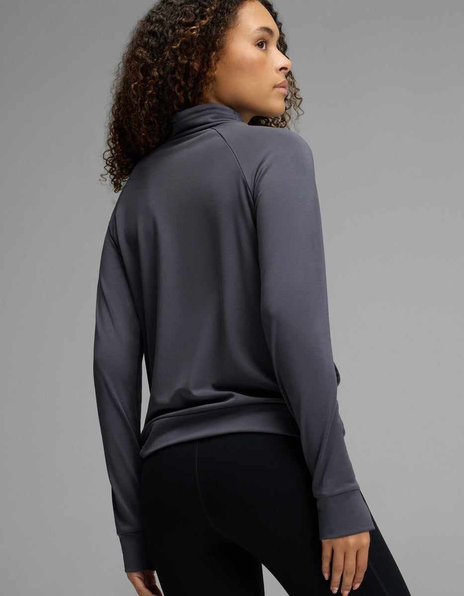 A person with curly hair wearing the Cozy Earth Women’s Brushed Bamboo Mock Neck in dark gray and black leggings stands facing away, looking over their shoulder against a plain gray background.