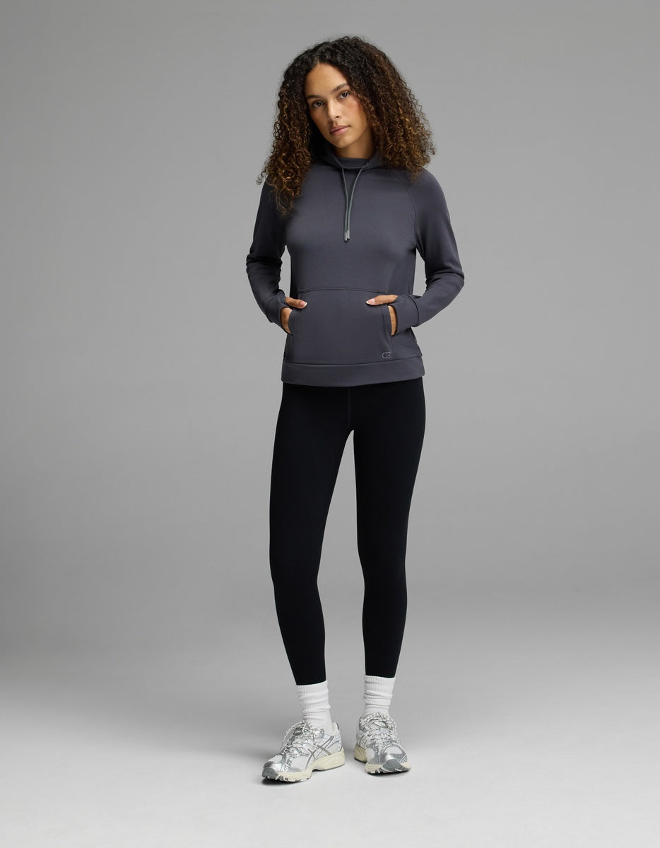 A person stands against a plain gray background, wearing the Cozy Earth Women’s Brushed Bamboo Mock Neck with their hands in the sweatshirt pockets, paired with black leggings, white socks, and white sneakers. Their curly hair falls loosely around their shoulders.
