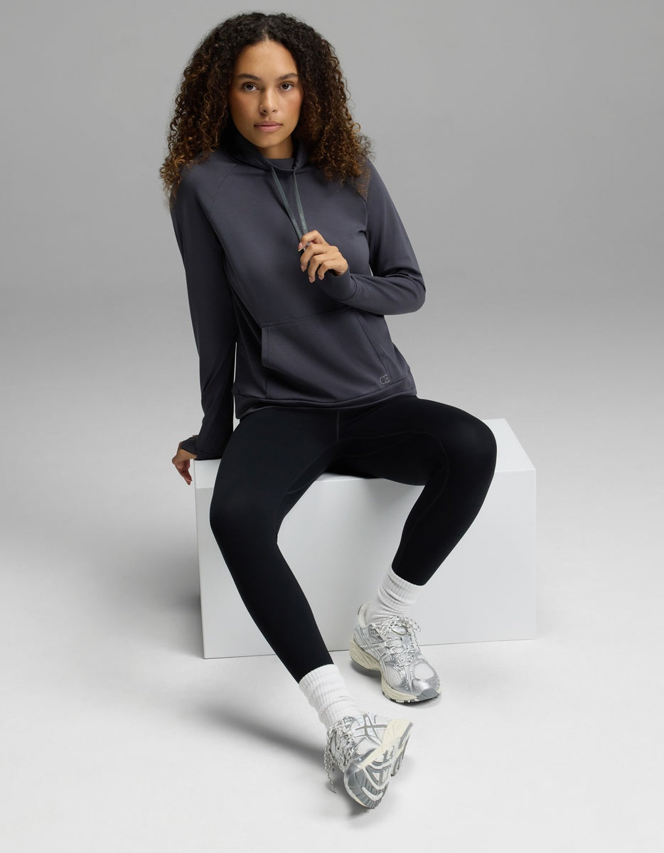 A person with curly hair wearing the Cozy Earth Women’s Brushed Bamboo Mock Neck, paired with black leggings and silver sneakers, sits on a white block in a studio with a plain gray background.
