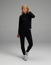A woman stands in a neutral pose against a plain gray background, wearing Cozy Earth’s Women’s Brushed Bamboo Mock Neck & Jogger Set in black, paired with white sneakers.|Color:Black