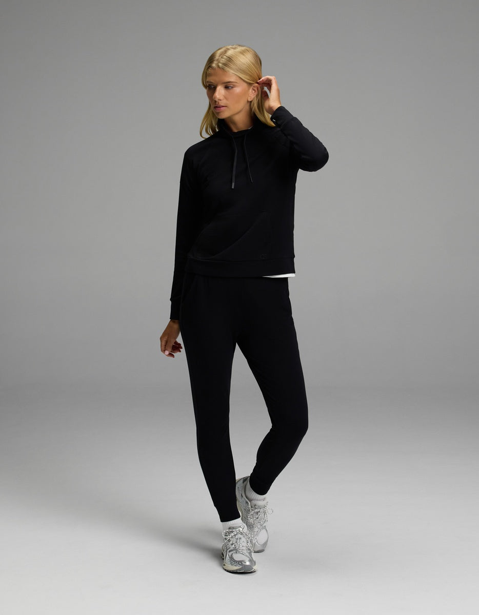 A woman stands in a neutral pose against a plain gray background, wearing Cozy Earth’s Women’s Brushed Bamboo Mock Neck & Jogger Set in black, paired with white sneakers.