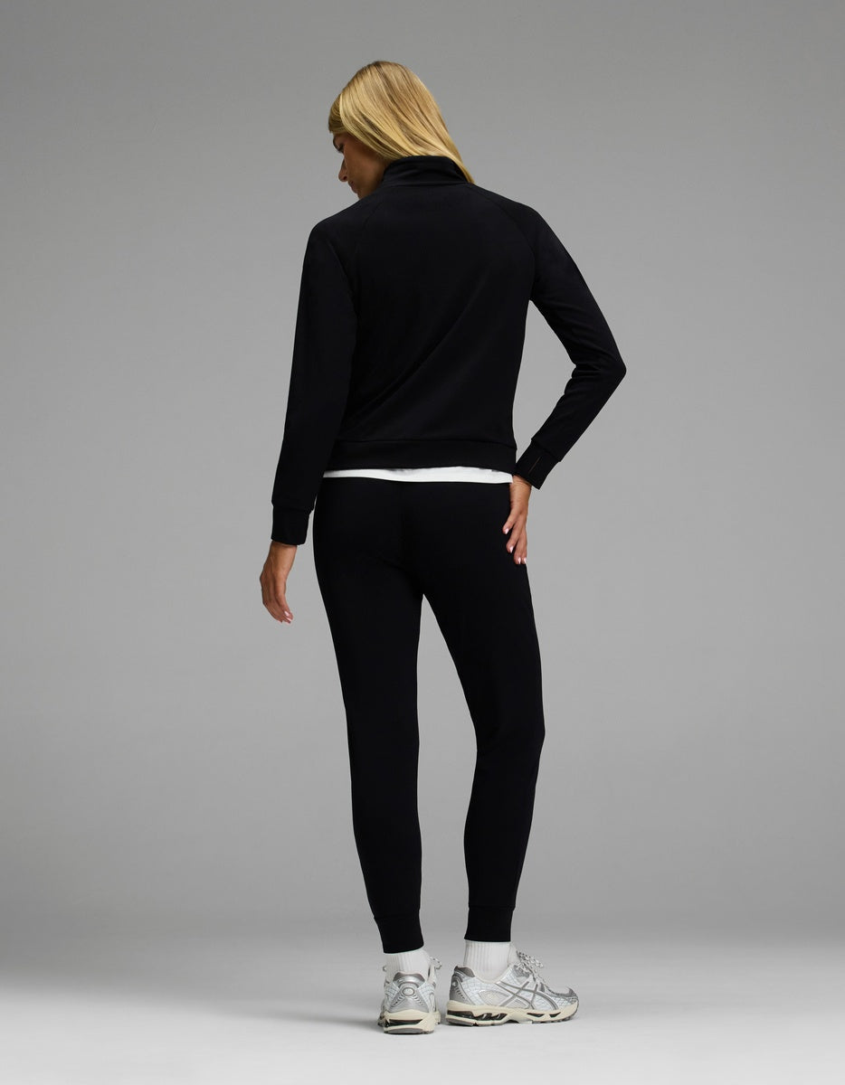 A person with long blonde hair stands facing away against a gray background, wearing the Cozy Earth Women’s Brushed Bamboo Mock Neck & Jogger Set in black, paired with a white shirt and gray sneakers.