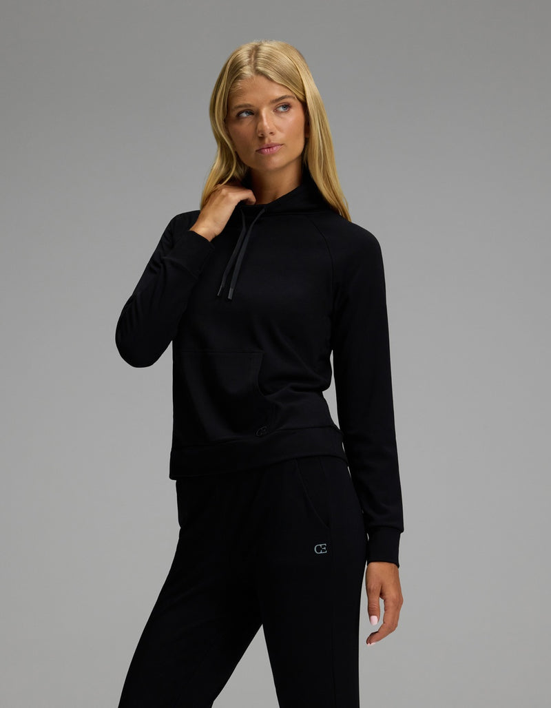 A woman with long blonde hair models the Cozy Earth Women’s Brushed Bamboo Mock Neck & Jogger Set in black, standing against a plain gray background with one hand near her collar and gazing slightly to the side.