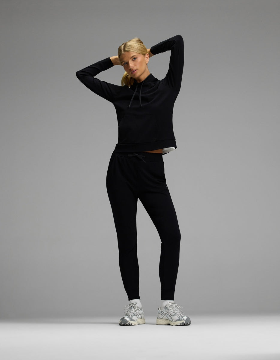 A person stands with hands behind their head, wearing the Cozy Earth Women’s Brushed Bamboo Mock Neck & Jogger Set in black, paired with white sneakers against a plain gray background.