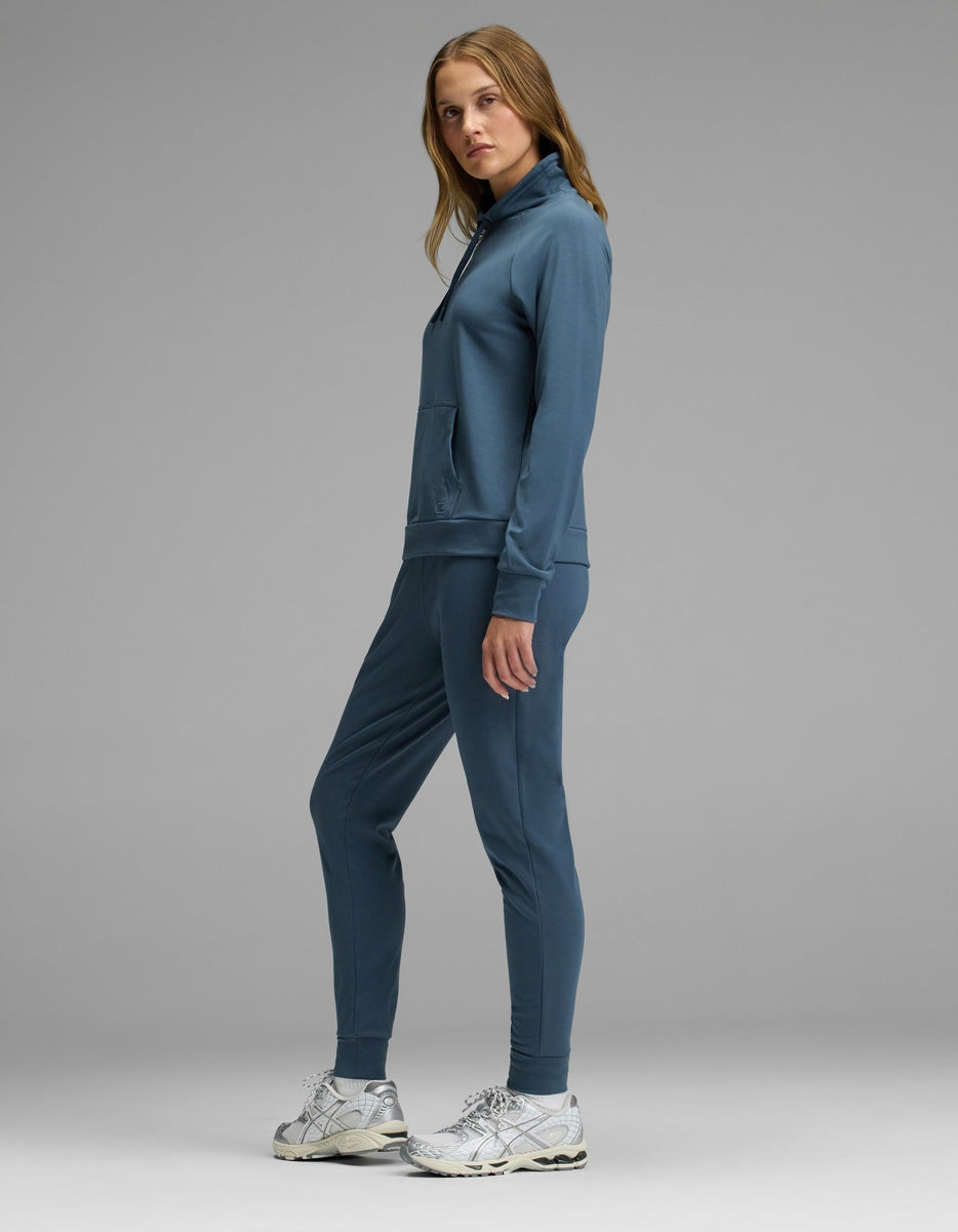 A woman stands sideways against a gray background, wearing Cozy Earth’s Women’s Brushed Bamboo Mock Neck with white sneakers. She looks at the camera with a neutral expression.|Color:Dawn
