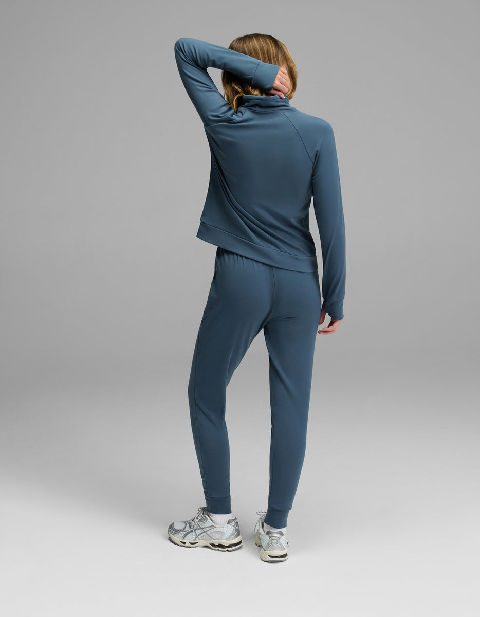 A person wears the Cozy Earth Women’s Brushed Bamboo Mock Neck & Jogger Set, paired with white sneakers, standing with one arm bent and covering part of their face, against a plain gray background.