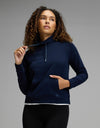 A woman with curly hair wears the Cozy Earth Women’s Brushed Bamboo Mock Neck in navy, paired with black leggings. She gazes upward and slightly to the side, holding a drawstring with one hand while her other hand rests in the front pocket, set against a neutral gray background.|Color:Navy