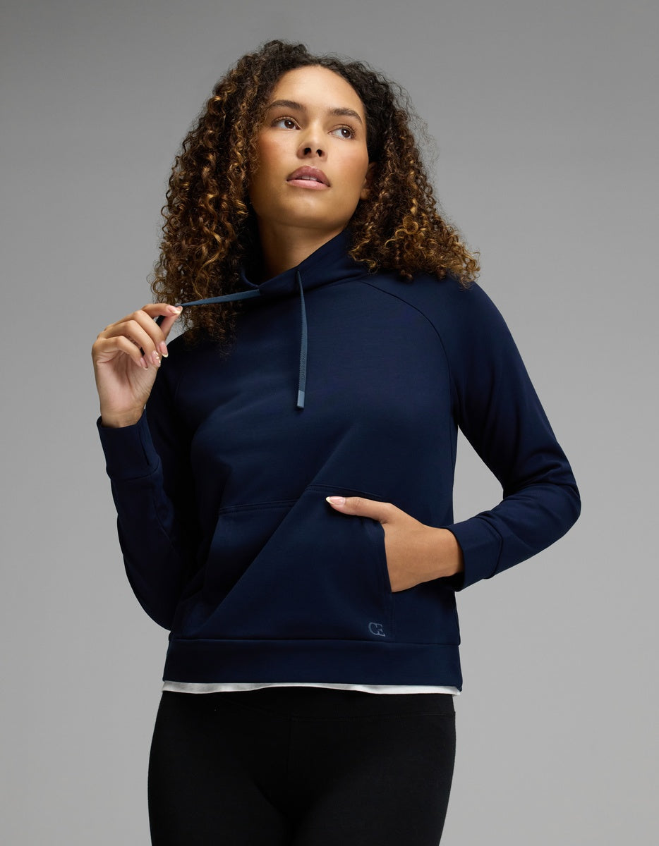 A woman with curly hair wears the Cozy Earth Women’s Brushed Bamboo Mock Neck in navy, paired with black leggings. She gazes upward and slightly to the side, holding a drawstring with one hand while her other hand rests in the front pocket, set against a neutral gray background.