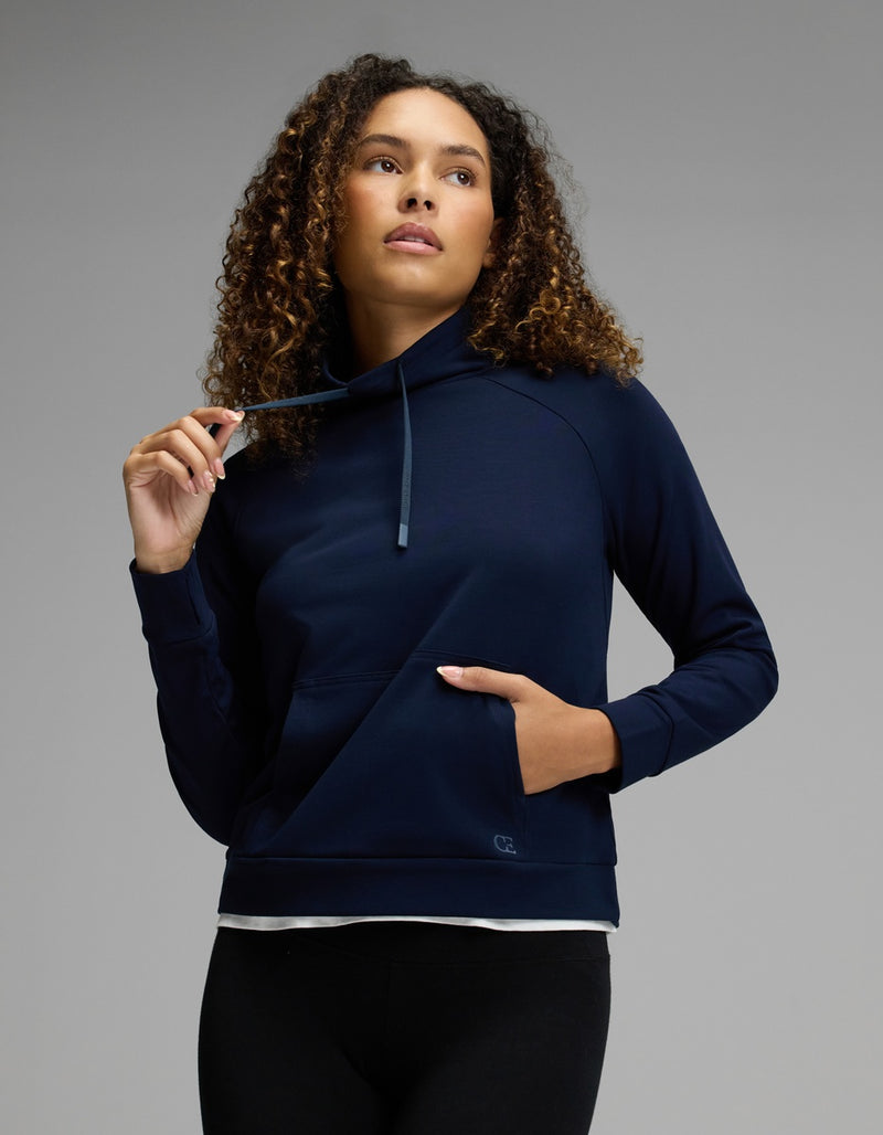 A woman with curly hair wears the Cozy Earth Women’s Brushed Bamboo Mock Neck in navy, paired with black leggings. She gazes upward and slightly to the side, holding a drawstring with one hand while her other hand rests in the front pocket, set against a neutral gray background.