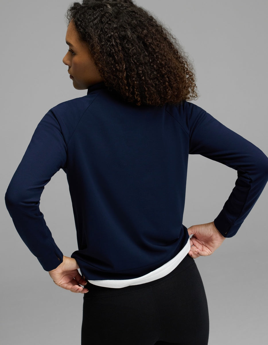 A woman with curly hair, wearing the Cozy Earth Women’s Brushed Bamboo Mock Neck in navy over a white shirt and black pants, stands with her back to the camera against a plain gray background.