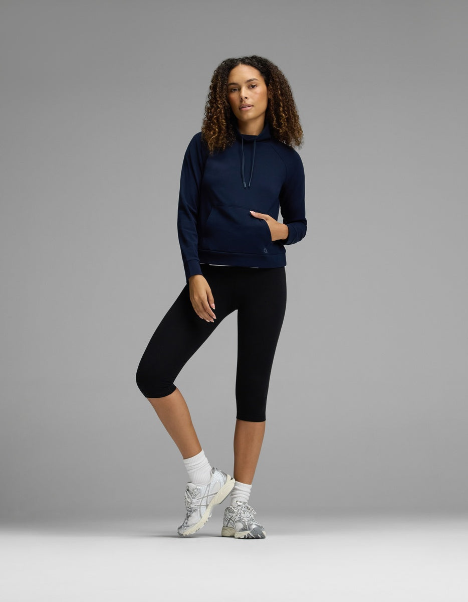 A woman with curly hair stands against a gray background with one leg slightly bent, wearing the Cozy Earth Women’s Brushed Bamboo Mock Neck in navy, paired with black capri leggings, white socks, and sneakers.