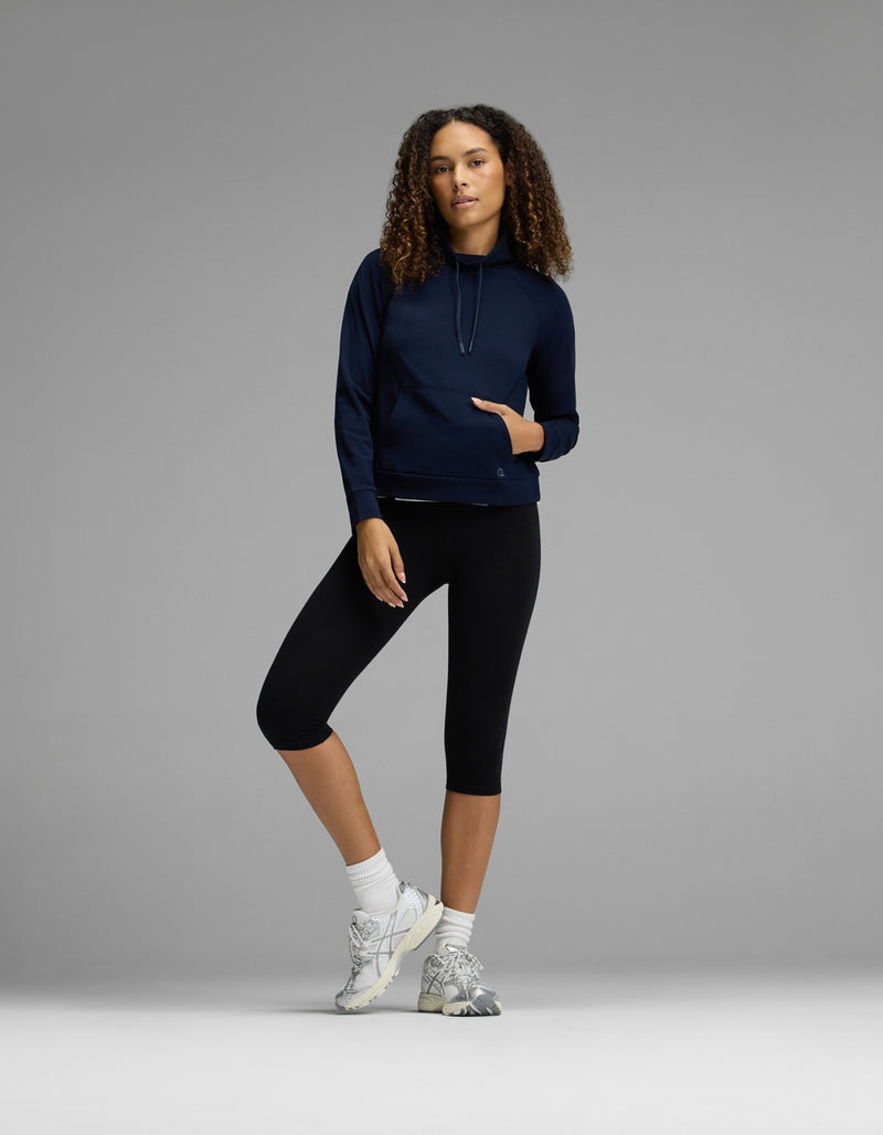 A woman with curly hair stands against a gray background with one leg slightly bent, wearing the Cozy Earth Women’s Brushed Bamboo Mock Neck in navy, paired with black capri leggings, white socks, and sneakers.
