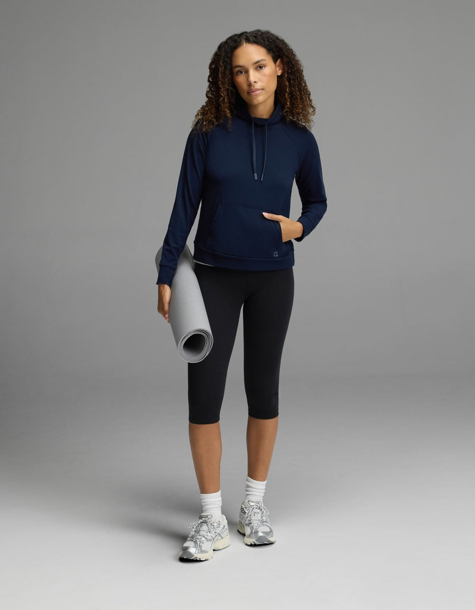 Wearing the Cozy Earth Women’s Brushed Bamboo Mock Neck in navy with black leggings, a person stands confidently against a gray background, holding a rolled-up yoga mat in one hand. They complete the look with white sneakers and have curly hair, gazing self-assuredly at the camera.
