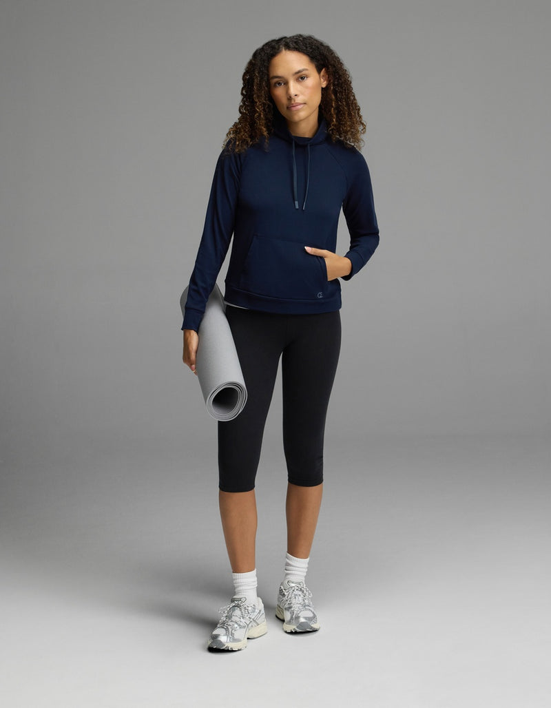 Wearing the Cozy Earth Women’s Brushed Bamboo Mock Neck in navy with black leggings, a person stands confidently against a gray background, holding a rolled-up yoga mat in one hand. They complete the look with white sneakers and have curly hair, gazing self-assuredly at the camera.