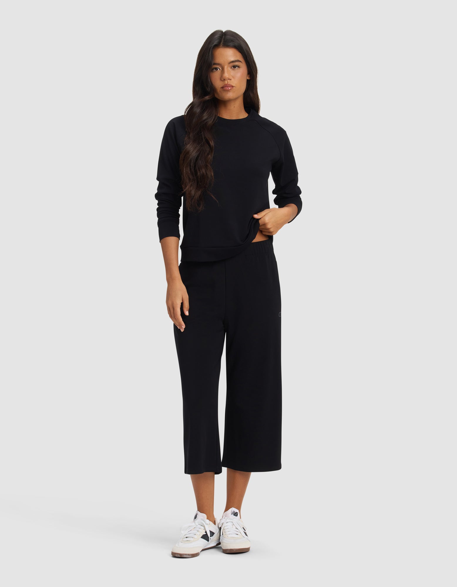 A woman with long dark hair wears Cozy Earth’s Women's Brushed Bamboo Pull On Capri in black, paired with a black long-sleeve sweatshirt and white sneakers, standing facing forward against a plain light gray background.