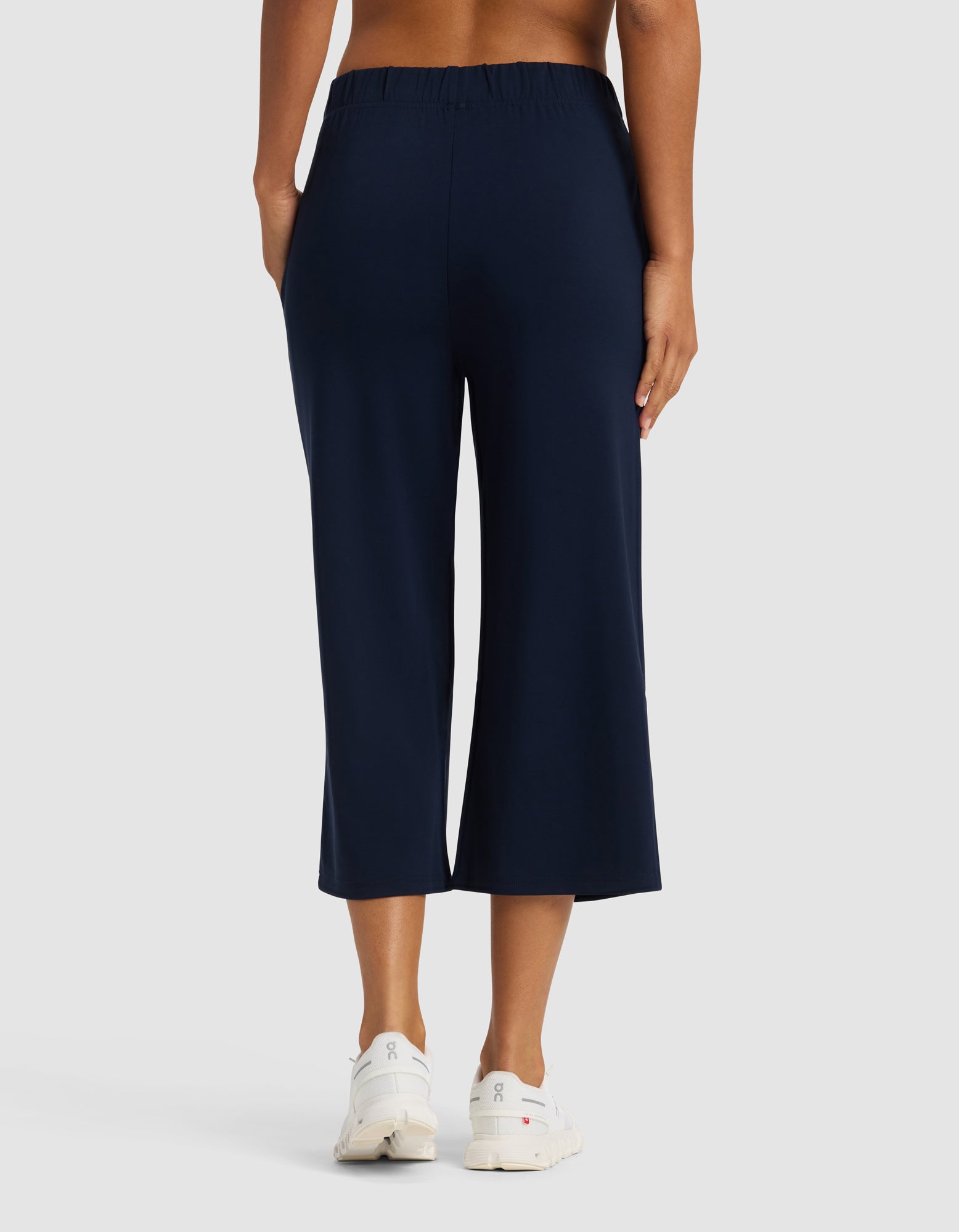A woman is seen from the back wearing Cozy Earth's Women's Brushed Bamboo Pull On Capri in navy blue with white sneakers, standing against a plain light gray background.|Color:Navy