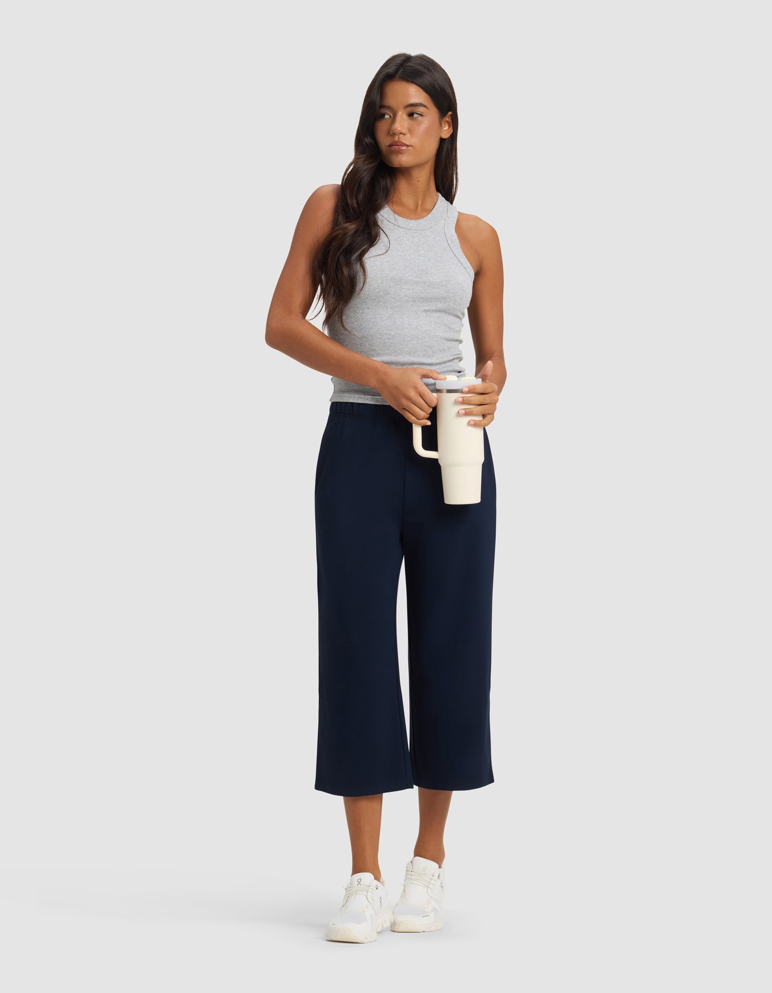 A woman with long dark hair wears Cozy Earth Women's Brushed Bamboo Pull On Capri, a gray sleeveless top, and white sneakers, holding a cream-colored travel mug against a plain light background.|Color:Navy