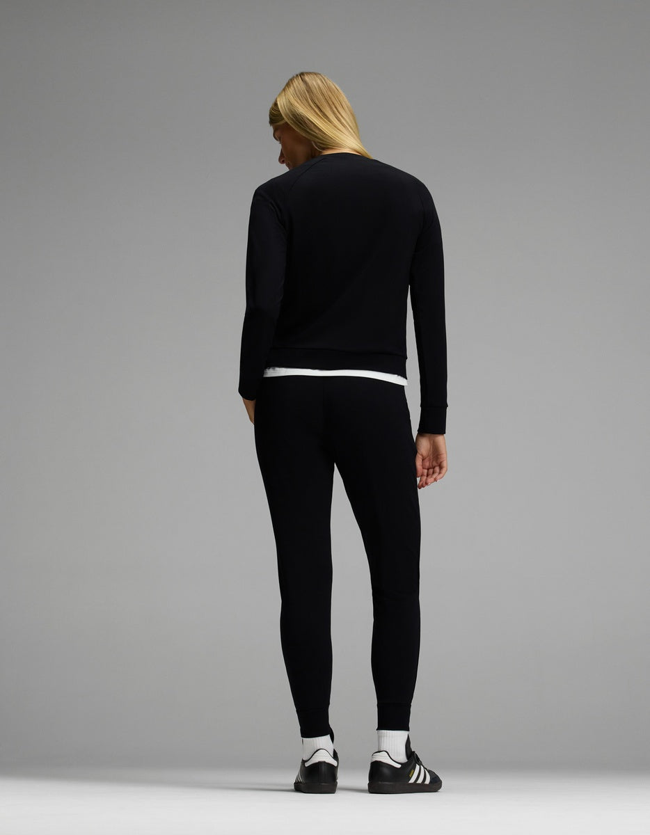 A person with long blond hair stands facing away, wearing the Cozy Earth Women’s Brushed Bamboo Jogger Set in black over a white undershirt and black and white Adidas sneakers against a plain light gray background.