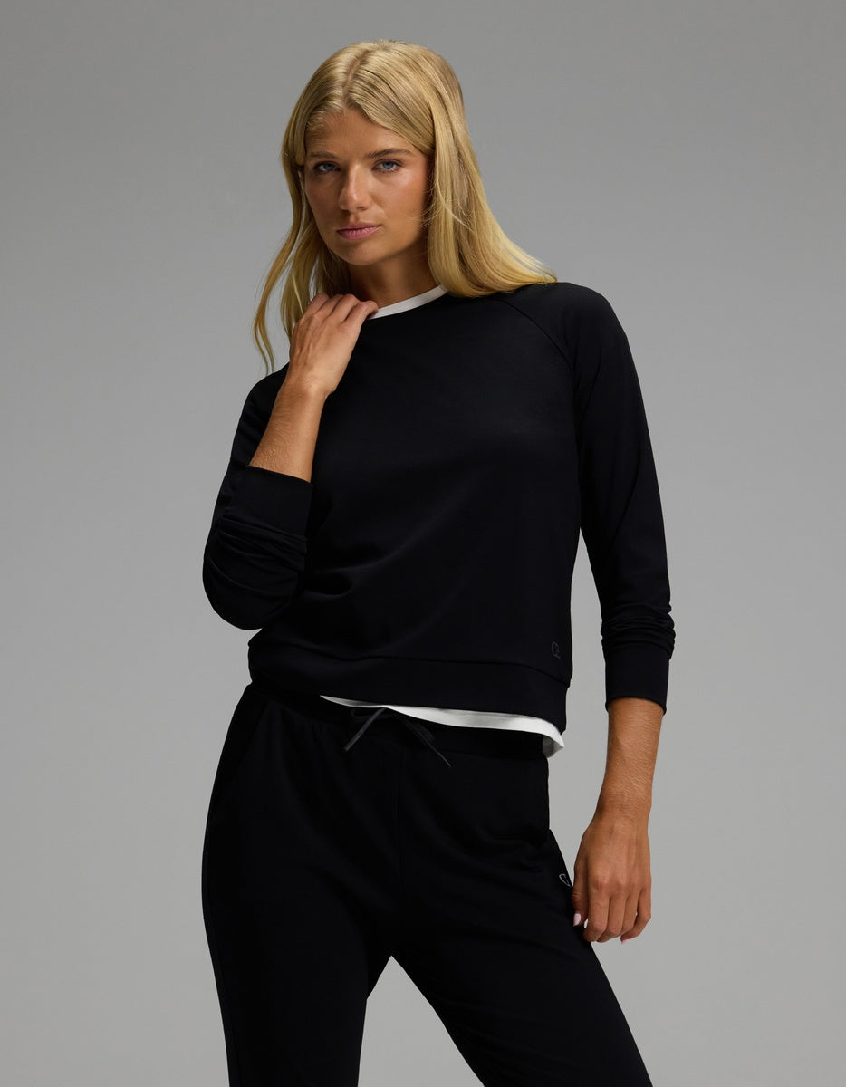 A woman with long blonde hair poses against a plain gray background, wearing the Cozy Earth Women’s Brushed Bamboo Jogger Set—featuring a black long-sleeve top layered over a white shirt and matching black pants.