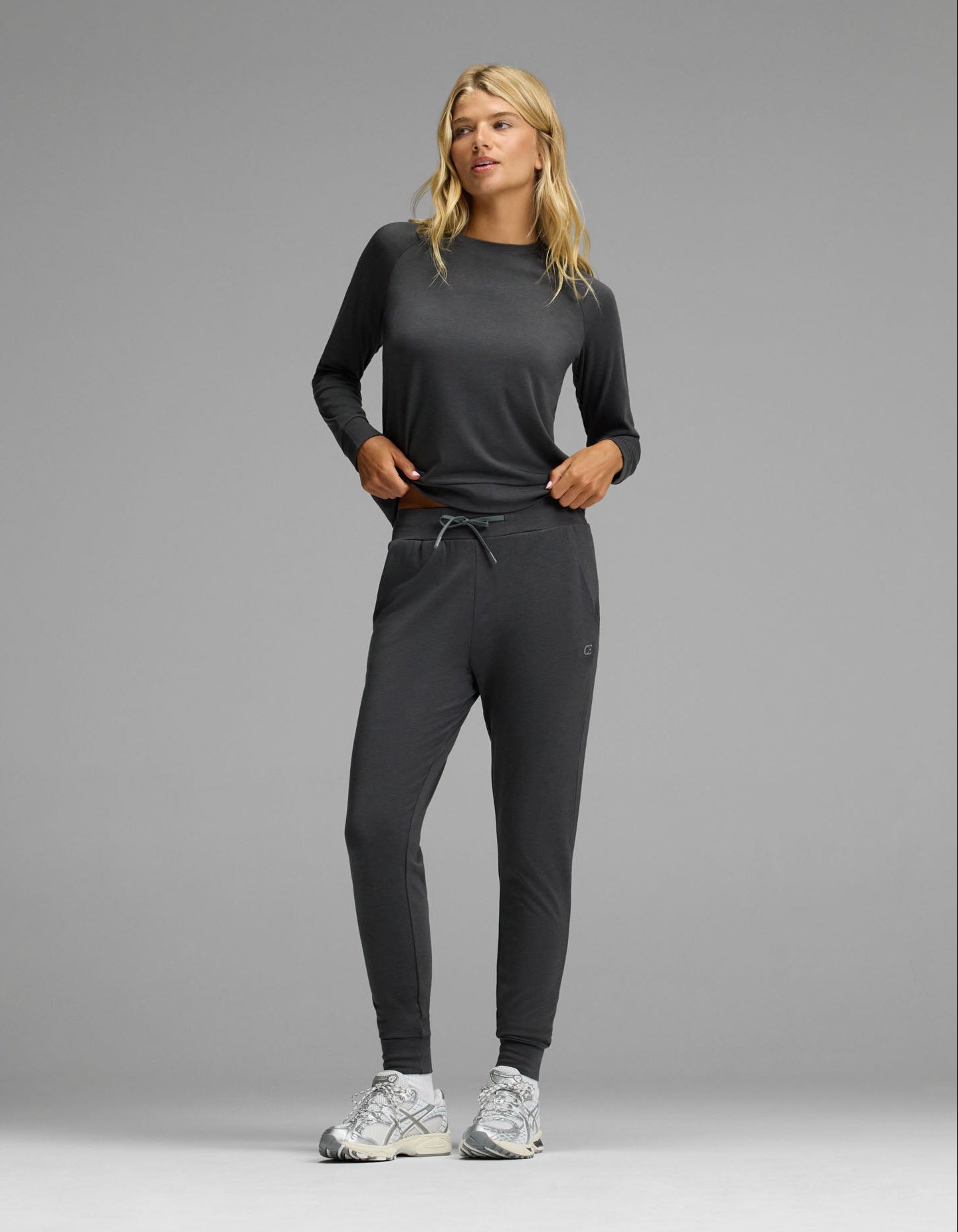 A woman with blonde hair stands against a gray background wearing the Cozy Earth Women’s Brushed Bamboo Jogger Set in Charcoal, paired with white sneakers. Her hands are in her pockets.|Color:Charcoal