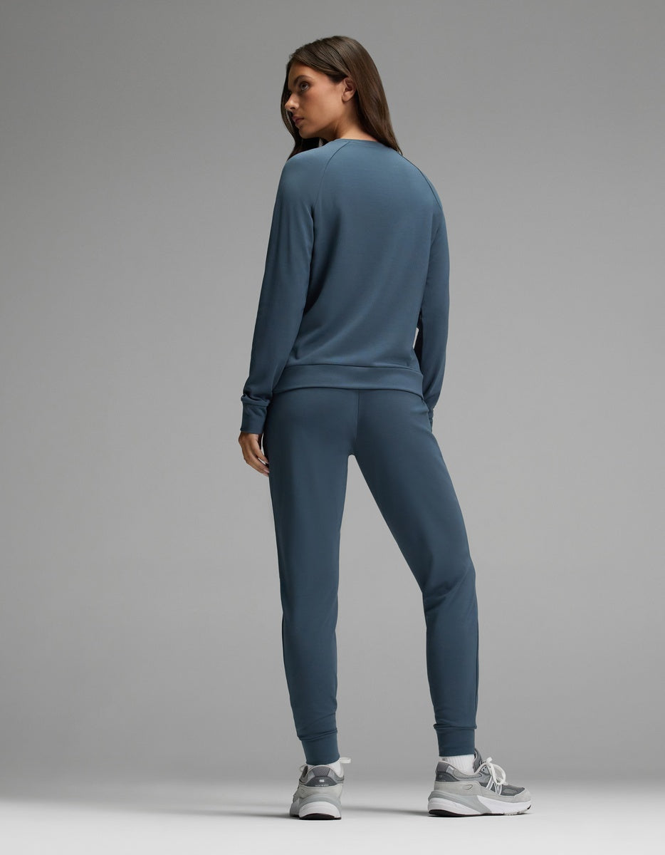 A woman stands facing away against a plain gray background, wearing the Cozy Earth Women’s Brushed Bamboo Jogger Set in blue, paired with gray sneakers.