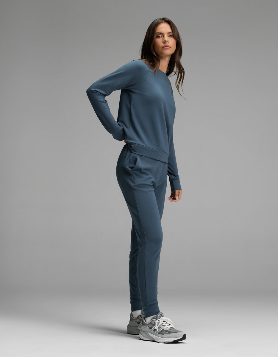 A woman stands in a studio wearing the Cozy Earth Women’s Brushed Bamboo Jogger Set, featuring a blue long-sleeve sweatshirt and matching jogger pants, paired with gray sneakers. She looks at the camera with a neutral expression against a plain gray background.
