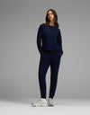 A woman models the Cozy Earth Women’s Brushed Bamboo Jogger Set in Navy, featuring a long-sleeve top and matching jogger pants with hands in the pockets, paired with white sneakers against a plain gray background.|Color:Navy