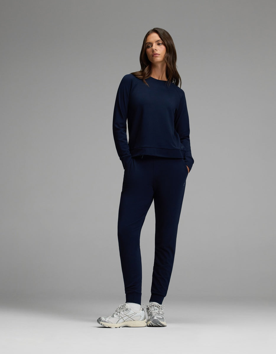 A woman models the Cozy Earth Women’s Brushed Bamboo Jogger Set in Navy, featuring a long-sleeve top and matching jogger pants with hands in the pockets, paired with white sneakers against a plain gray background.