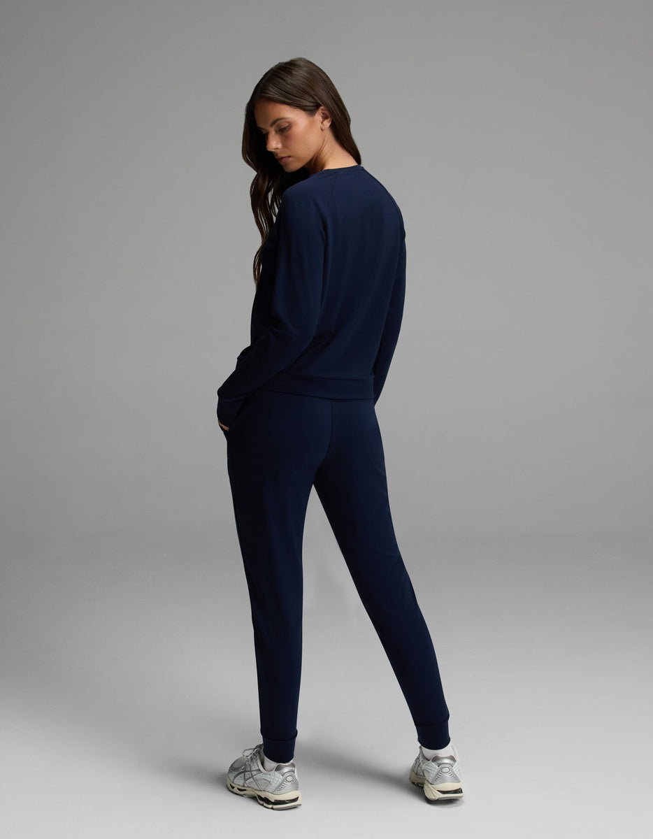 A woman stands facing away, looking over her shoulder. She is wearing the Cozy Earth Women’s Brushed Bamboo Jogger Set in navy blue, paired with white sneakers. The background is plain and gray.