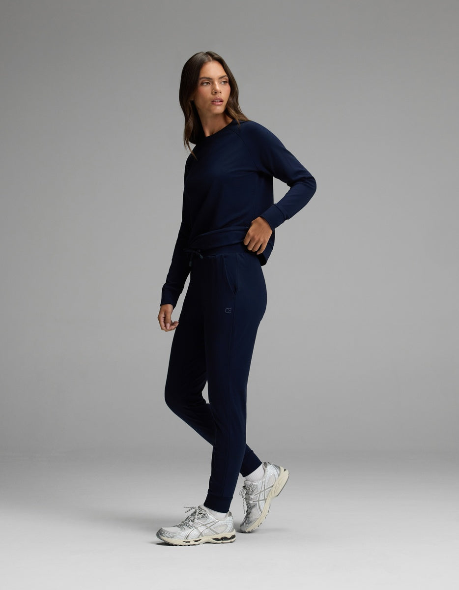 A woman wearing the Cozy Earth Women’s Brushed Bamboo Jogger Set in navy blue with white sneakers stands against a plain gray background.