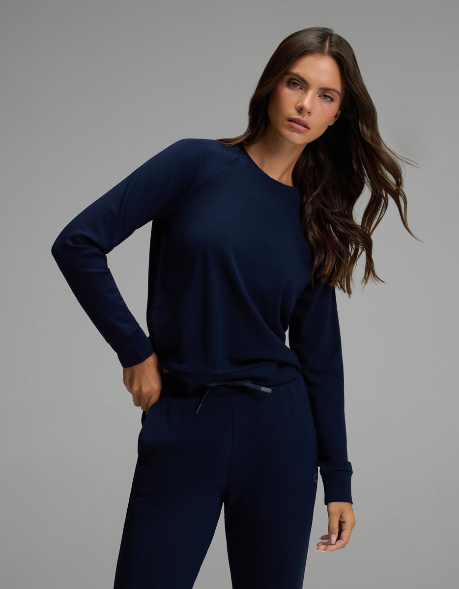 A woman with long brown hair wears the Cozy Earth Women’s Brushed Bamboo Jogger Set—a matching navy blue, long-sleeve top and jogger pants. She stands against a plain gray background, one hand in her pocket and a neutral expression on her face.