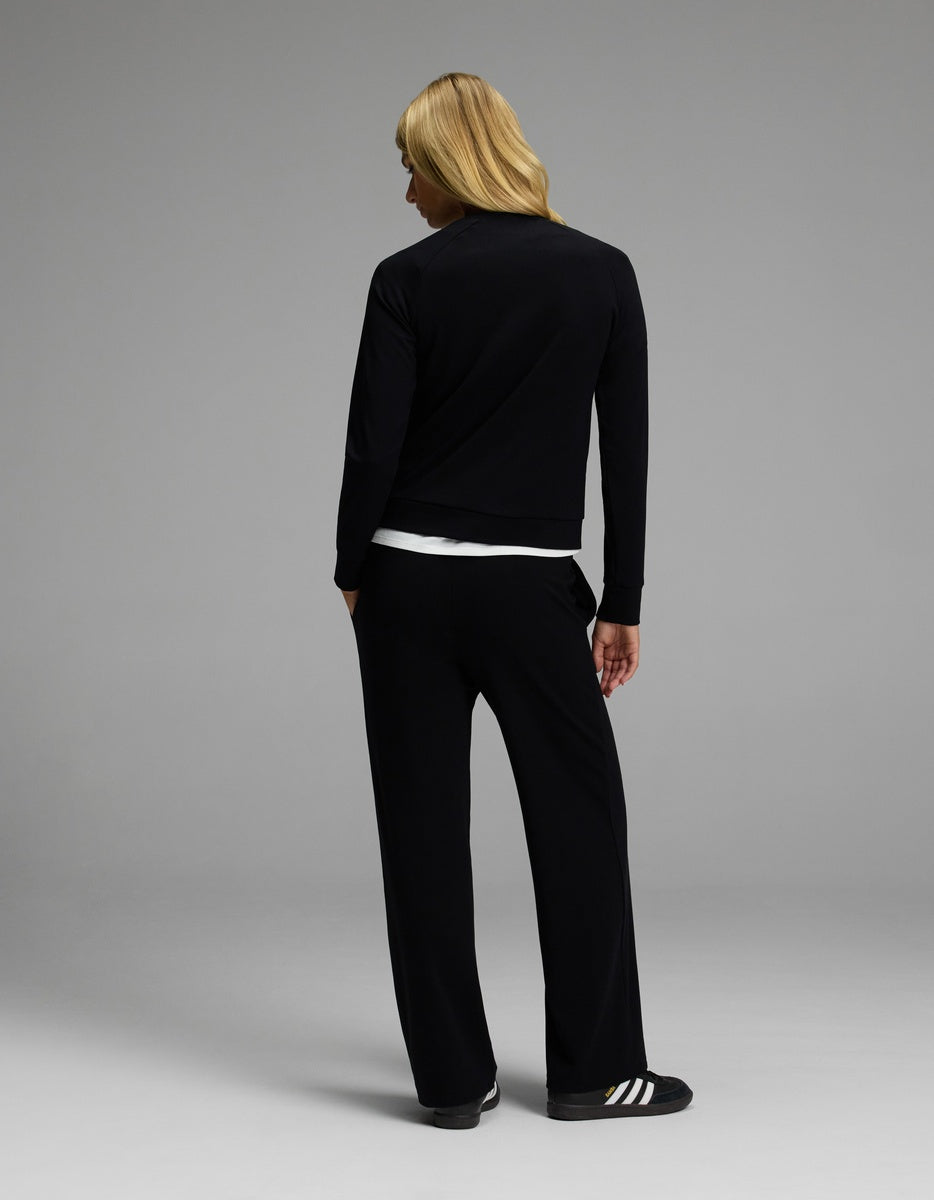 A person with long blond hair stands facing away, dressed in the Cozy Earth Women’s Brushed Bamboo Wide Leg Pull On Pant & Pullover Crew Set in black, with a white shirt visible underneath. The background is plain and gray.