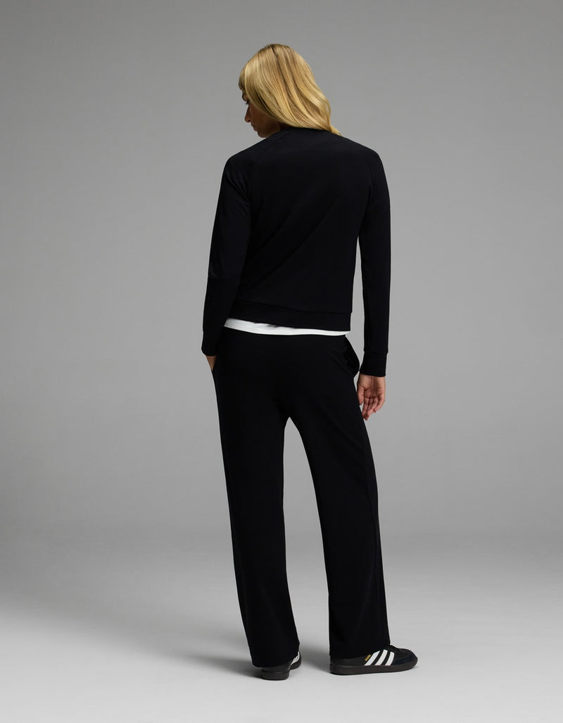 A person with long blond hair stands facing away, dressed in the Cozy Earth Women’s Brushed Bamboo Wide Leg Pull On Pant & Pullover Crew Set in black, with a white shirt visible underneath. The background is plain and gray.