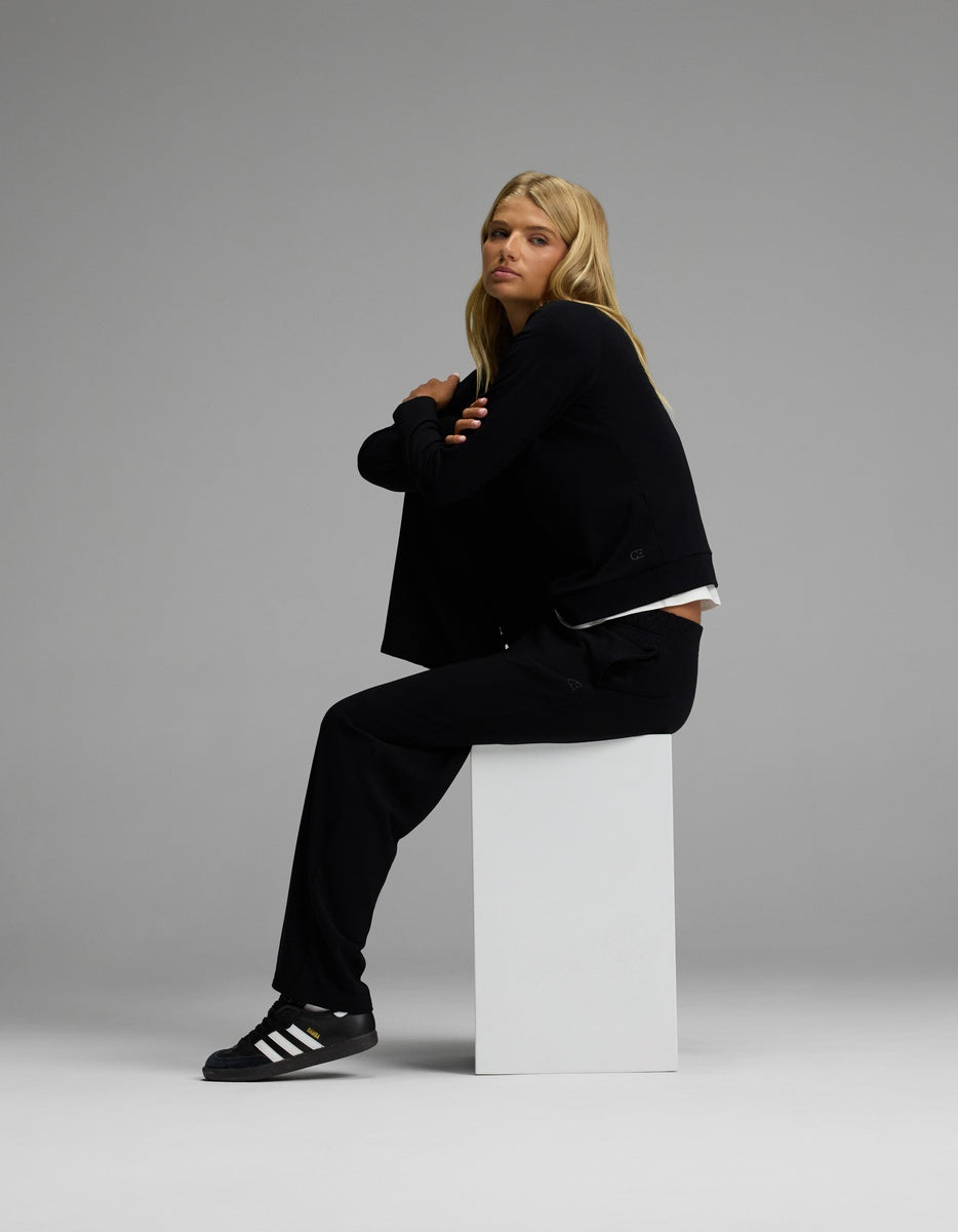 A person with long blonde hair sits sideways on a white block, wearing the Women’s Brushed Bamboo Wide Leg Pull On Pant & Pullover Crew Set from Cozy Earth, set against a plain gray background.