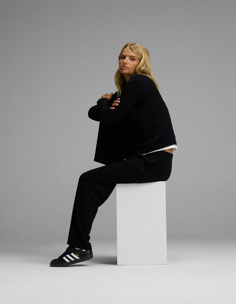 A person with long blonde hair sits sideways on a white block, wearing the Women’s Brushed Bamboo Wide Leg Pull On Pant & Pullover Crew Set from Cozy Earth, set against a plain gray background.