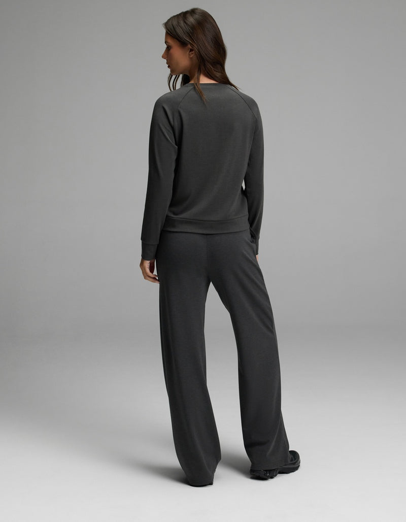 A woman with long brown hair stands facing away, wearing the Cozy Earth Women’s Brushed Bamboo Wide Leg Pull On Pant & Pullover Crew Set in dark gray against a plain light gray background.