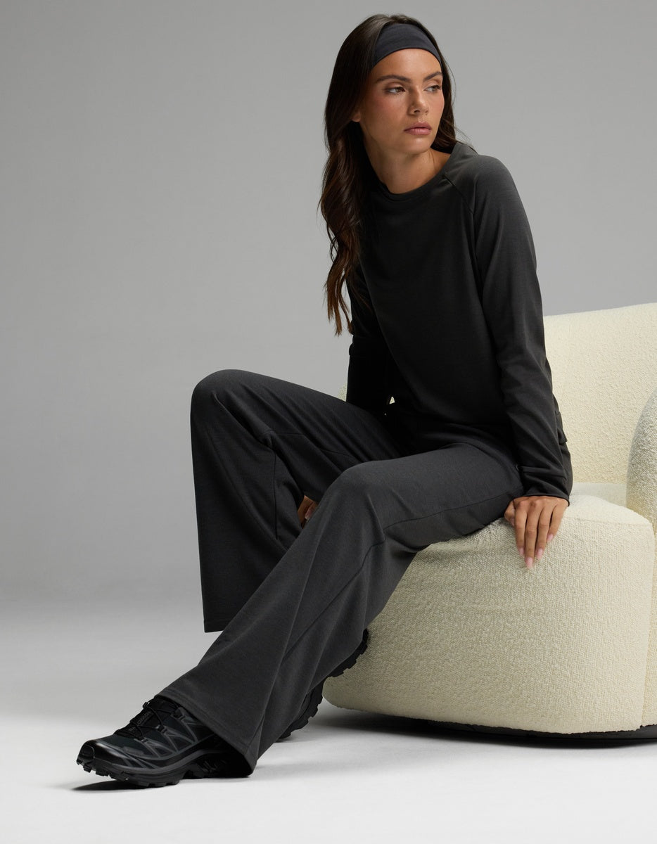 A woman models the Cozy Earth Women’s Brushed Bamboo Wide Leg Pull On Pant & Pullover Crew Set in a dark hue, paired with black shoes and a matching headband. She sits on a cream-colored chair, gazing to the side against a plain gray background.