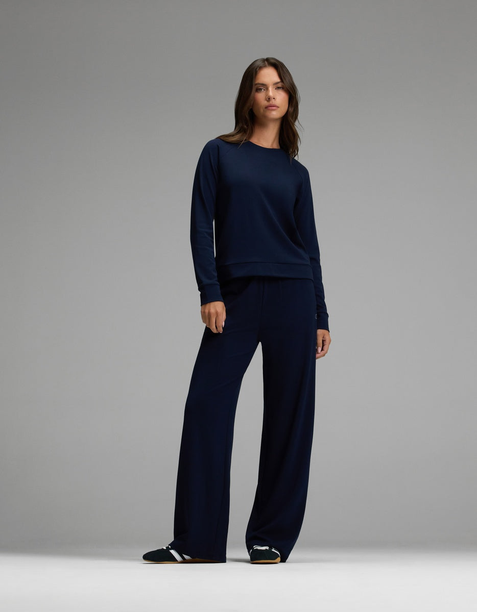 A woman stands against a gray background wearing the Cozy Earth Women’s Brushed Bamboo Wide Leg Pull On Pant & Pullover Crew Set in navy, paired with black slide sandals.