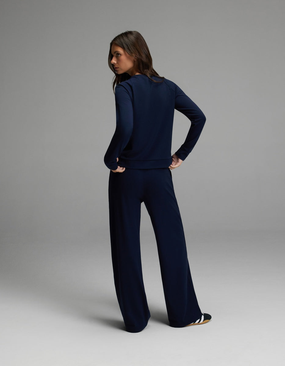 A woman stands facing away with her hands on her hips, wearing the Cozy Earth Women’s Brushed Bamboo Wide Leg Pull On Pant & Pullover Crew Set in navy. The background is a simple, light gray studio setting.