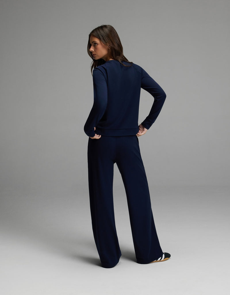 A woman stands facing away with her hands on her hips, wearing the Cozy Earth Women’s Brushed Bamboo Wide Leg Pull On Pant & Pullover Crew Set in navy. The background is a simple, light gray studio setting.