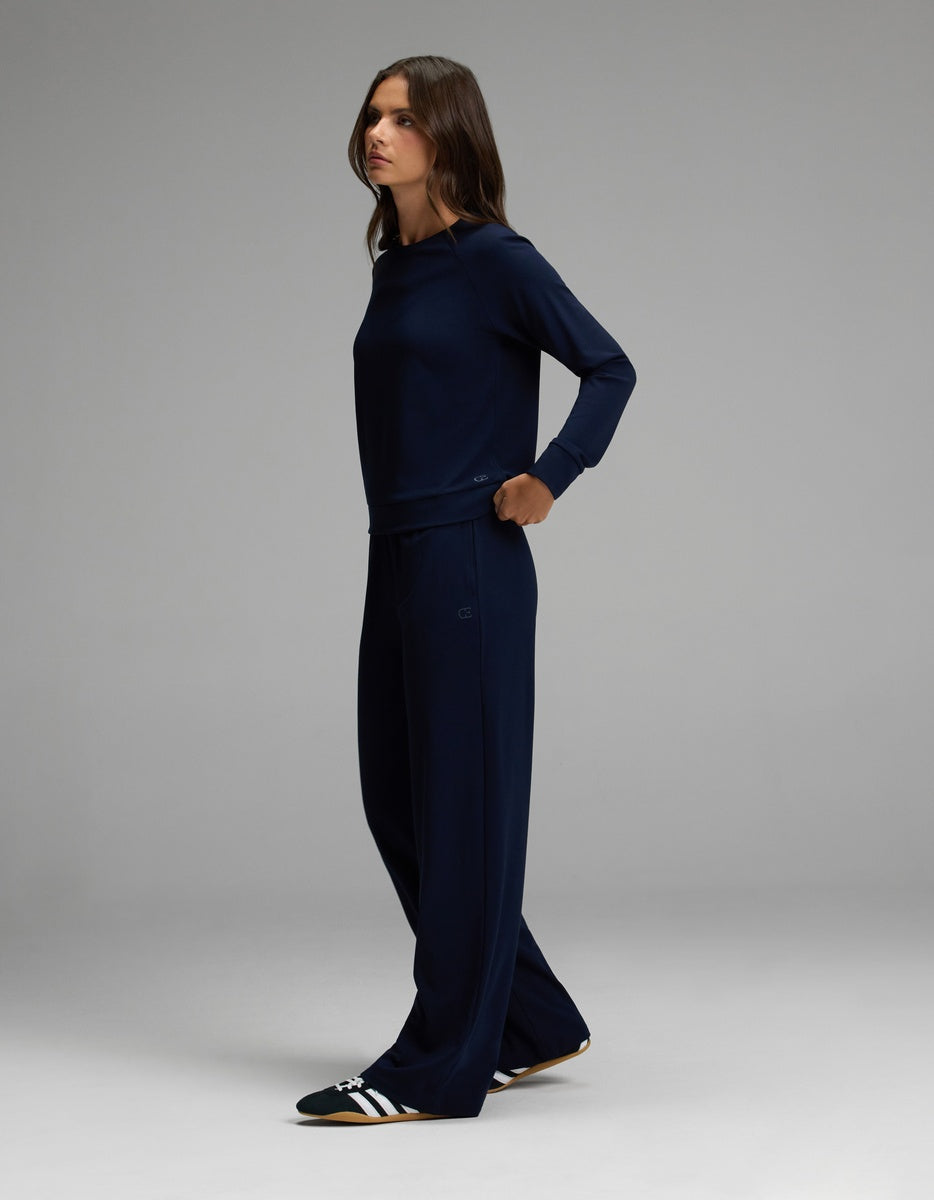 A woman stands sideways against a plain gray background, wearing the Cozy Earth Women’s Brushed Bamboo Wide Leg Pull On Pant & Pullover Crew Set in dark blue, paired with black and white slide sandals.