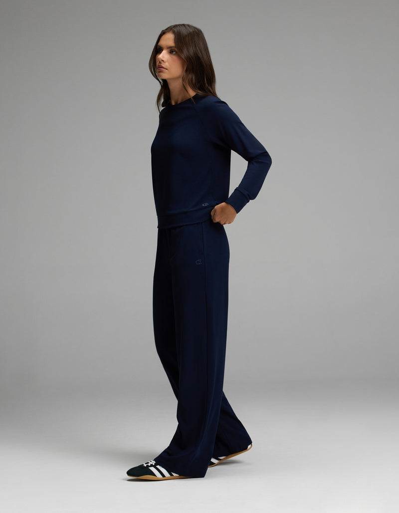 A woman stands sideways against a plain gray background, wearing the Cozy Earth Women’s Brushed Bamboo Wide Leg Pull On Pant & Pullover Crew Set in dark blue, paired with black and white slide sandals.