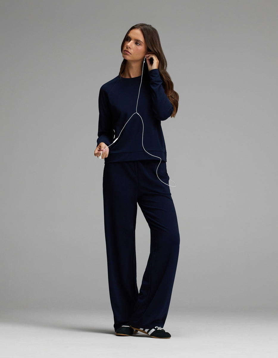 A woman wearing the Cozy Earth Women’s Brushed Bamboo Wide Leg Pull On Pant & Pullover Crew Set in navy and striped slippers stands against a gray background, listening to music with wired earphones.